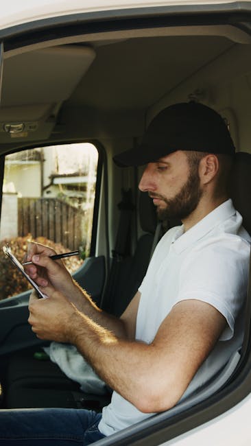 A man with a beard wearing a black cap and white t-shirt is seated inside the cab of a delivery van, holding a clipboard and pen in his hands. The interior of the vehicle shows a beige ceiling and side panels, with a window revealing a garden and wooden fence outside. The man appears to be reviewing or noting information related to the home relocation process. The van is positioned on a paved driveway, and the background includes blurred foliage. This scene illustrates a professional moving service, such as those provided by [COMPANY_NAME], involved in packing, loading, or coordinating furniture transport during a house removal.