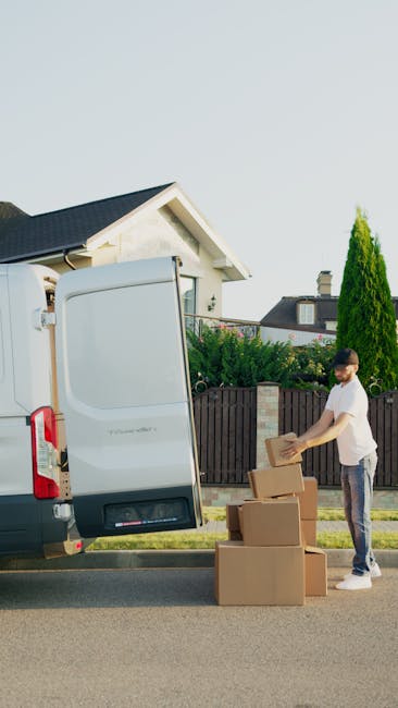 A man with a beard, wearing a white t-shirt and jeans, is standing on the pavement next to a large white moving van with its rear doors open. He is stacking or unpacking brown cardboard boxes, which are placed on the ground in front of him. The boxes are of various sizes and some appear to be sealed with packing tape. Inside the van, visible through the open doors, there are more cardboard boxes and packaging materials. The scene is set in a residential area with a house, a wooden fence, and greenery including a tall coniferous tree in the background. The bright daylight and clear sky suggest a sunny day, and the surroundings indicate the process of home relocation involving furniture transport and packing, typical of a professional removals service like Man With a Van Belmont.