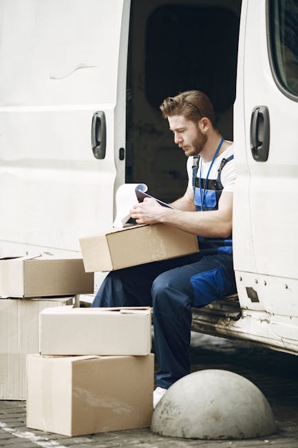 A man with a beard wearing a black cap and white t-shirt is seated inside the cab of a delivery van, holding a clipboard and pen in his hands. The interior of the vehicle shows a beige ceiling and side panels, with a window revealing a garden and wooden fence outside. The man appears to be reviewing or noting information related to the home relocation process. The van is positioned on a paved driveway, and the background includes blurred foliage. This scene illustrates a professional moving service, such as those provided by [COMPANY_NAME], involved in packing, loading, or coordinating furniture transport during a house removal.