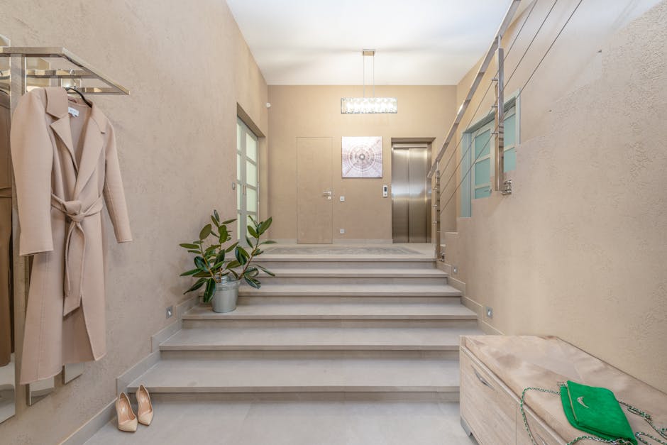 Image of a residential lobby area showing beige tiled stairs leading to an upper level, with a potted green plant placed on one of the stairs. To the left, a beige coat is hanging on a wall-mounted rack, and a pair of beige high-heeled shoes are positioned on the floor nearby. The wall on the left side features a frosted glass door, while the back wall has a modern, rectangular ceiling light fixture and a framed decorative artwork. On the right side, a metal and glass railing runs along the staircase, providing safety. At the bottom right corner, there is a closed wooden chest with a green handbag and chain strap resting on top. The environment appears clean and well-lit, suitable for house removals or home relocation activities, with items that could be part of packing and moving processes handled by [COMPANY_NAME].