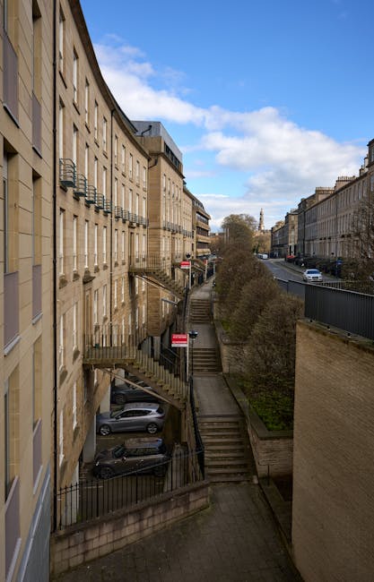 View of a multi-storey residential building with external staircases leading down to a lower level, where several parked cars are visible in a small parking area. The image captures a pathway with steps and metal railing, situated between the building and a section of greenery with bushes. On the street adjacent to the building, a grey van is parked, indicating possible loading or unloading activity related to house removals or home relocation services. The scene is under a partly cloudy sky with natural daylight illuminating the surroundings. This image highlights the logistics of moving furniture and boxes through staircases and narrow pathways typical of urban properties, as observed by Man With a Van Belmont, specialists in removals and relocation solutions, showcasing an appropriate environment for furniture transport and packing and moving processes.
