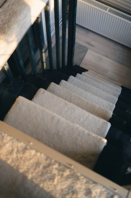 Image of a residential lobby area showing beige tiled stairs leading to an upper level, with a potted green plant placed on one of the stairs. To the left, a beige coat is hanging on a wall-mounted rack, and a pair of beige high-heeled shoes are positioned on the floor nearby. The wall on the left side features a frosted glass door, while the back wall has a modern, rectangular ceiling light fixture and a framed decorative artwork. On the right side, a metal and glass railing runs along the staircase, providing safety. At the bottom right corner, there is a closed wooden chest with a green handbag and chain strap resting on top. The environment appears clean and well-lit, suitable for house removals or home relocation activities, with items that could be part of packing and moving processes handled by [COMPANY_NAME].