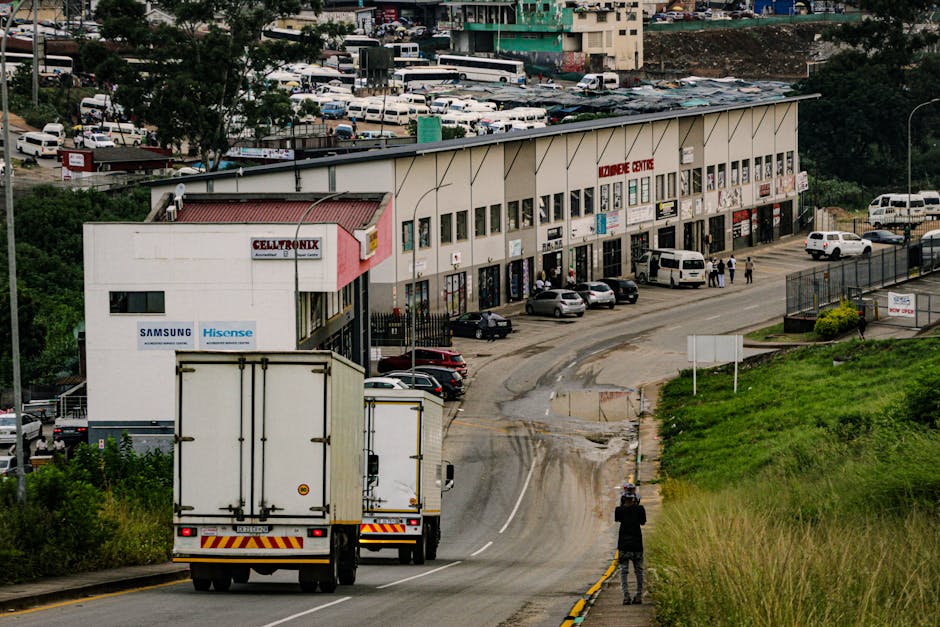 A white moving truck with rear doors closed is driving along a narrow, slightly winding urban street with visible tire marks and patches of mud on the asphalt, indicating ongoing or recent vehicle movement. Behind the truck, a white van is visible in front of a large, multi-level commercial building labeled 'Belmont Centre,' which has multiple shopfronts with signage, including 'Celltronix' and brand advertisements for Samsung and Hisense. Several cars are parked along the front of the building, with some people walking near the entrances, possibly engaged in shopping or visiting the premises. To the right of the street, a grassy embankment with green vegetation slopes uphill, and a person dressed in dark clothing is standing on the sidewalk, capturing the scene with a camera or phone. The overall environment suggests a busy local area with a combination of commercial activity and ongoing home or business relocations, with [COMPANY_NAME] likely involved in the logistics of furniture transport and packing during a move.