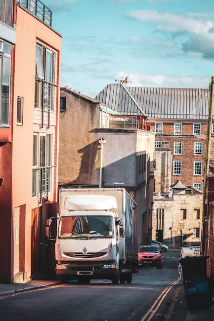 A white box truck from Man With a Van Belmont is parked on a narrow street adjacent to multi-storey residential buildings with a mix of brick and rendered facades in a city area. The truck is positioned parallel to the pavement, with its cab facing towards the camera, and appears to be in the process of a home relocation or furniture transport. Nearby, there are several cardboard boxes and wrapped furniture items loaded onto or ready for loading into the vehicle, with some boxes stacked on the pavement close to the truck. The street features a slight incline, with other vehicles parked further along the road, including a black car and a smaller red vehicle. The scene is illuminated by daylight, with shadows cast by the buildings indicating a clear sky. This setting depicts the logistics involved in house removals, with items being prepared or loaded for transport as part of a professional moving service, consistent with the services offered by Man With a Van Belmont in the Belmont Road area.