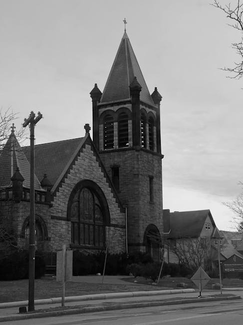 A black and white photograph of a large church building featuring a prominent tall, pointed steeple with a weather vane at the top, situated among other smaller structures in the background. The church's facade is made of stone with arched windows and decorative brickwork. To the left, a utility pole with multiple wires is visible, and the church is adjacent to a paved street with a curb and road signs. The scene appears to be in a quiet residential or suburban area, possibly during overcast weather, with some trees visible without leaves in the surroundings. This image may be used in a moving guide to illustrate transportation or logistical considerations for relocating properties near historic or notable buildings, such as the church, which might impact access or parking during a home relocation or furniture transport process. Man With a Van Belmont offers removals services that include careful handling of such properties during moving and packing activities.