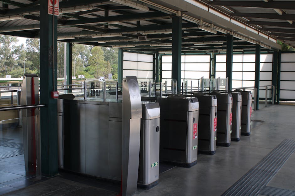 Inside an outdoor parking area of a transportation hub, a row of stainless steel turnstiles with black bases are positioned along a tiled floor. The turnstiles are equipped with card readers and contactless access points, used for entry and exit control. Above, a metal roof with exposed beams provides shelter, supported by green-painted metal columns. Transparent glass panels surround the open sides of the structure, allowing visibility of lush trees and a roadway with vehicles in the background, indicating a suburban or semi-rural setting. The area is clean and orderly, suitable for vehicle loading or passenger movement during house removals or relocation processes. The overall appearance reflects a modern, functional space designed for efficient flow of people and vehicles, aligning with the logistics involved in furniture transport and home relocation. Man With a Van Belmont’s presence is implied through context, emphasizing the focus on moving services and access management during a house move.