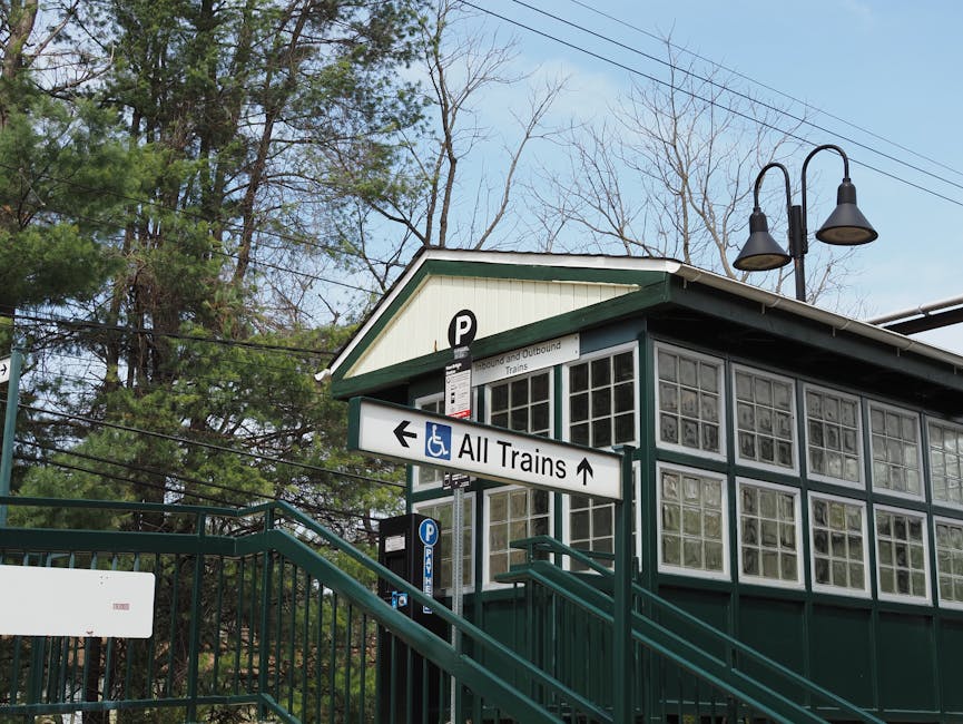 A black and white photograph of a large church building featuring a prominent tall, pointed steeple with a weather vane at the top, situated among other smaller structures in the background. The church's facade is made of stone with arched windows and decorative brickwork. To the left, a utility pole with multiple wires is visible, and the church is adjacent to a paved street with a curb and road signs. The scene appears to be in a quiet residential or suburban area, possibly during overcast weather, with some trees visible without leaves in the surroundings. This image may be used in a moving guide to illustrate transportation or logistical considerations for relocating properties near historic or notable buildings, such as the church, which might impact access or parking during a home relocation or furniture transport process. Man With a Van Belmont offers removals services that include careful handling of such properties during moving and packing activities.