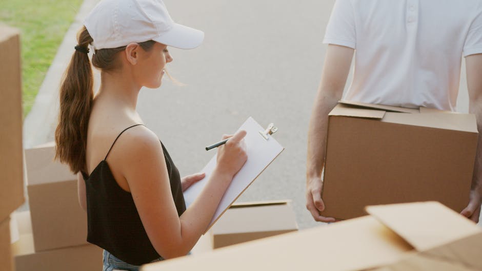 Close-up view of a person wearing dark clothing, handling a medium-sized cardboard box secured with packing tape. The individual is standing inside a property, near the doorway, with their hands gripp