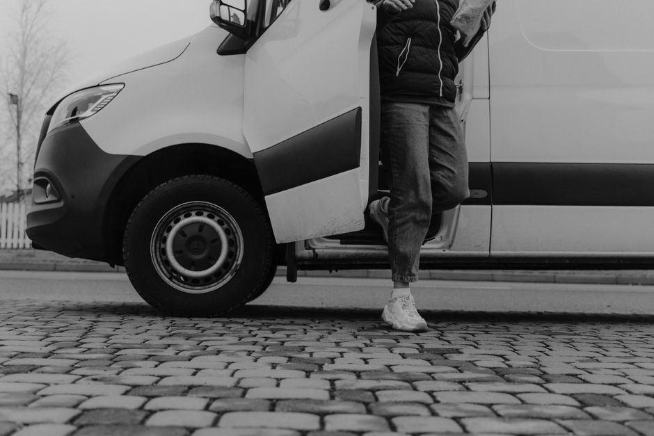 A black Ford Transit van, with a sleek and modern design, is parked on a paved driveway outside a contemporary office building with large glass windows and a connecting sky bridge. The van's front gri