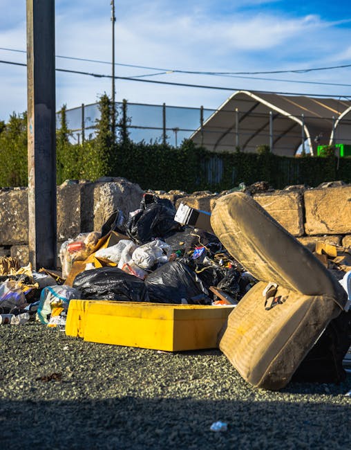 An outdoor scene showing a pile of bulky waste and household rubbish, including black plastic trash bags, cardboard boxes, and a large discarded beige upholstered armchair with visible fabric wear, placed on a gravel surface next to a stone boundary wall. A yellow plastic container is positioned in front of the waste, with cardboard packaging nearby. In the background, a wooden fence, utility poles with power lines, and a partially visible greenhouse or canopy structure are seen under a blue sky with scattered clouds. The setting indicates a collection point for waste removal, fitting with house removals and furniture transport services provided by Man With a Van Belmont, which manages logistics for packing, loading, and disposal during home relocation processes.