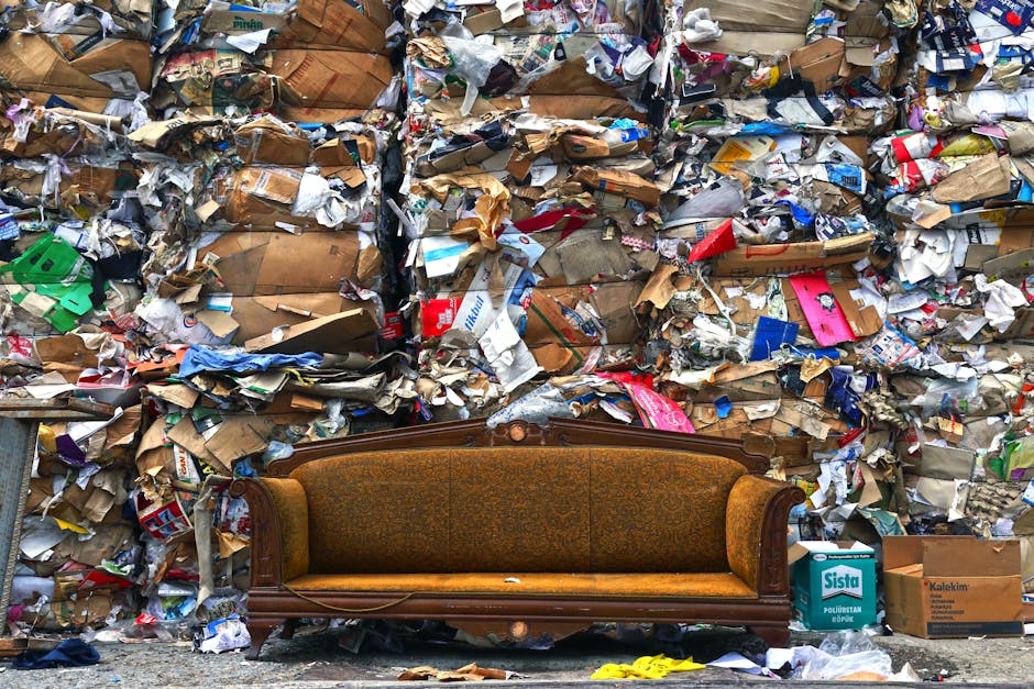 An outdoor scene showing a pile of bulky waste and household rubbish, including black plastic trash bags, cardboard boxes, and a large discarded beige upholstered armchair with visible fabric wear, placed on a gravel surface next to a stone boundary wall. A yellow plastic container is positioned in front of the waste, with cardboard packaging nearby. In the background, a wooden fence, utility poles with power lines, and a partially visible greenhouse or canopy structure are seen under a blue sky with scattered clouds. The setting indicates a collection point for waste removal, fitting with house removals and furniture transport services provided by Man With a Van Belmont, which manages logistics for packing, loading, and disposal during home relocation processes.