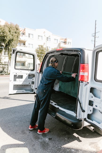A person dressed in dark clothing and red shoes is loading or unloading a large, green item wrapped in protective material into the spacious cargo area of a white van with open side and rear doors. Th