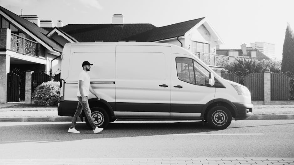 A person dressed in dark clothing and red shoes is loading or unloading a large, green item wrapped in protective material into the spacious cargo area of a white van with open side and rear doors. Th