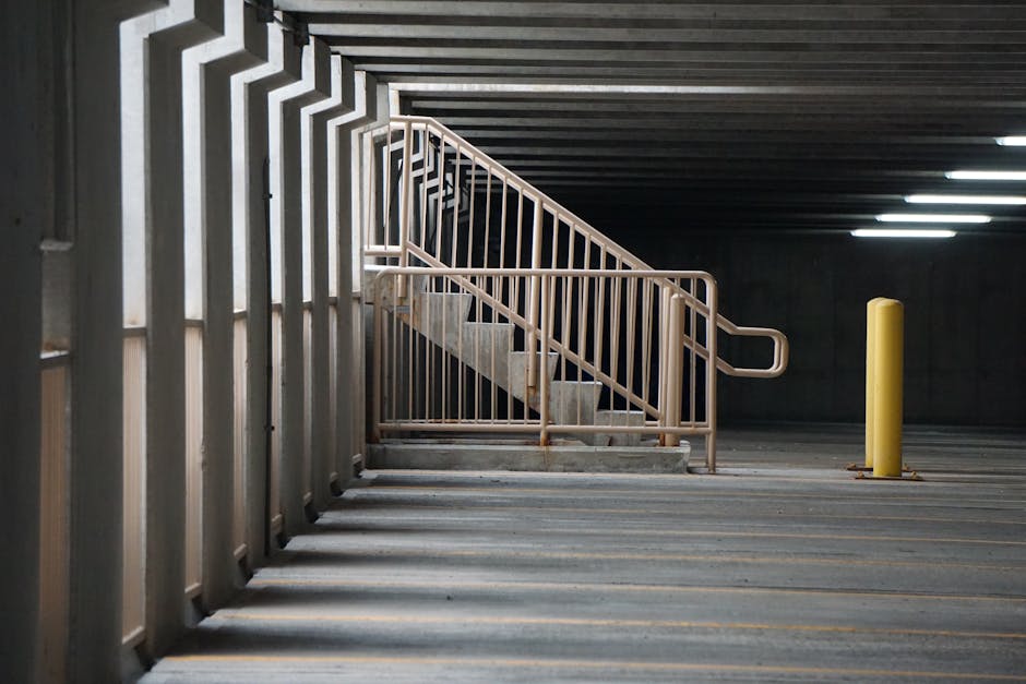 An indoor parking garage with a concrete staircase featuring a beige metal handrail that slopes upward to a higher level. The staircase is flanked by vertical metal bars on the side, integrated into a fencing structure. The parking area has a dark ceiling with embedded fluorescent lighting, illuminating the space evenly. To the right of the staircase, a yellow protective bollard is positioned on the concrete floor, which has faint yellow parking lines painted across it. The scene is clean and empty, capturing the environment typical of a residential or commercial building's basement parking area, relevant to home relocation and furniture transport logistics handled by Man With a Van Belmont.