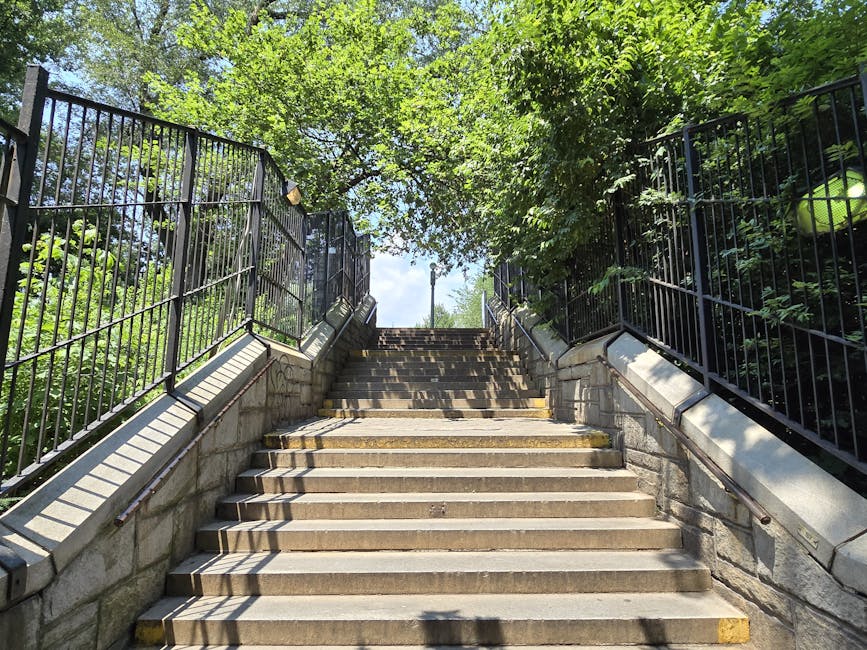 A set of outdoor concrete stairs leading up to a residential area, with black metal railings on both sides. The staircase is surrounded by leafy green trees and shrubs, casting dappled shadows on the steps. The steps are wide and uniform, with some showing slight wear from use. At the top of the stairs, there is a clear sky visible through the foliage, and a lamp post can be seen in the distance. The scene suggests an outdoor home relocation environment, with the staircase part of a property accessed during furniture transport or packing and moving activities. The surrounding greenery and the open sky indicate it is daytime, and the overall setting is calm and typical of a suburban or park-adjacent residential area, which [COMPANY_NAME] may encounter during house removals or moving services, emphasizing the need for careful navigation and logistics planning for delivery, stairs, parking, and accessibility considerations.