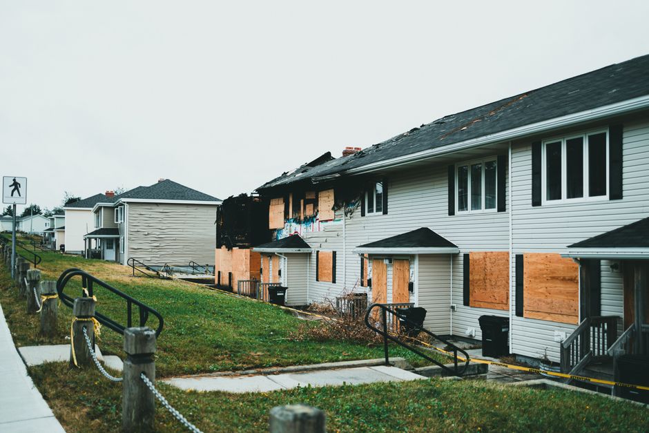 The image shows a row of residential houses with a large section of the exterior wall damaged by fire, exposing burnt and blackened interior walls, with some windows and doors boarded up with plywood. The damaged house is situated on a paved sidewalk with a grass verge alongside, featuring metal railings and concrete posts connected by chains, separating the pavement from the properties. To the left, a pedestrian crossing sign is visible, indicating proximity to a pedestrian crossing area. The neighbouring houses appear intact, with clean facades and no visible damage. The sky is overcast, contributing to a neutral and subdued lighting environment. This scene depicts a home undergoing repair or assessment, relevant to house removals or property damage inspection services provided by companies such as Man With a Van Belmont, emphasizing the importance of secure property handling and safe logistics during moving processes.