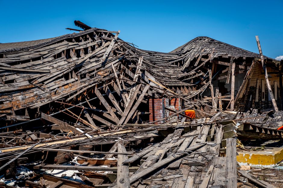 A large section of a collapsed wooden roof structure shows broken and splintered beams, some of which are still attached to brick chimneys, amidst scattered debris including broken wood, roofing materials, and collapsed parts of the building's facade, all visible under a clear blue sky. The damage likely resulted from a severe structural failure, with the damaged roof hanging in irregular, jagged formations. Nearby, a portion of the concrete pavement and a yellow-painted curb are visible, indicating the location is at the edge of a property or street. This scene is indicative of extensive damage at a residential or commercial property, with the debris and fallen materials occupying both interior and exterior spaces. The image captures a moment in a home or property after destruction, which could be relevant to property removal or insurance assessment, illustrating the extent of structural failure and the importance of careful handling during moving or recovery processes, as undertaken by companies like Man With a Van Belmont.