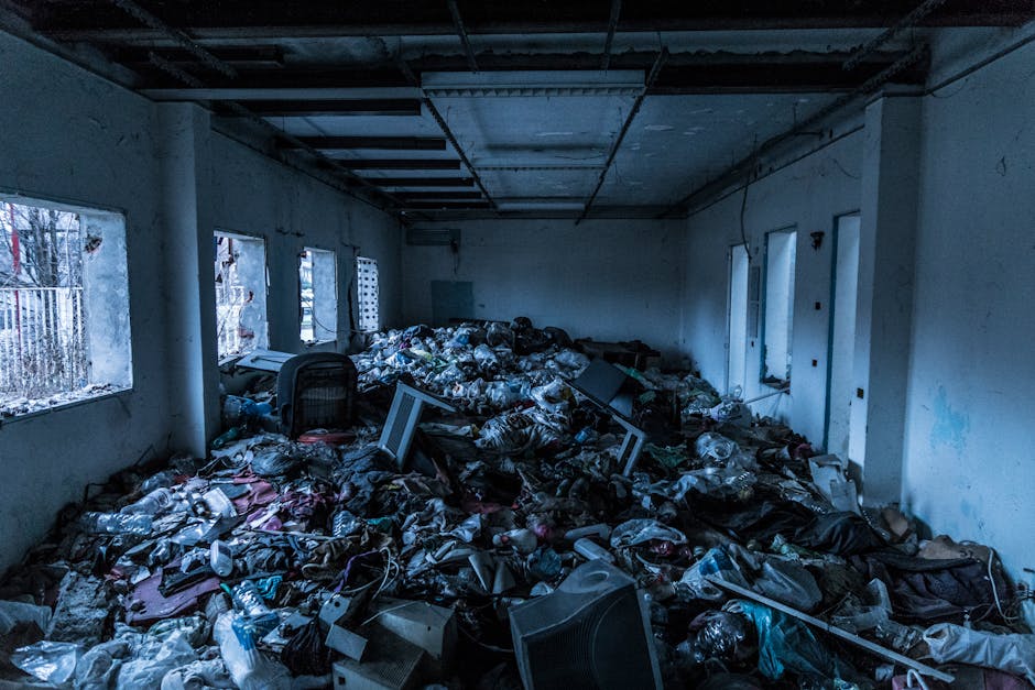 An indoor scene showing a room filled with a large accumulation of household waste, including plastic bottles, paper, cardboard boxes, and discarded electronic items scattered across the floor. The room has four windows along one wall, allowing natural light to illuminate the space, which appears damp and unclean with partly painted walls. In the foreground, an office chair is partially buried beneath the clutter. In the background, the room's ceiling shows exposed beams and ceiling panels, indicating it is under renovation or in disrepair. A team from Man With a Van Belmont is managing the clear-up process, with visible effort in removing and sorting the debris, preparing the space for a comprehensive house clearance as part of a property renovation or relocation project. The scene reflects the initial stages of packing and waste removal involved in a professional furniture transport or home relocation service.