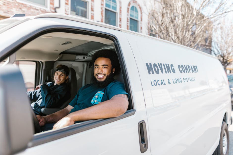 Inside a white moving van labeled 'Moving Company Local & Long Distance,' two men are visible preparing for a home relocation. One man with a beard and dark curly hair is seated at the driver's seat, smiling at the camera, while the second man, with straight dark hair and wearing a headband, is seated in the passenger seat, also smiling. The van's interior shows a glimpse of packing materials and equipment, with the open side door revealing part of the loading process. Outside the van, nearby residential buildings and bare trees indicate an outdoor setting typical for house removals, with natural daylight illuminating the scene. This image depicts professional furniture transport and packing activities associated with residential moves, aligned with services offered by Man With a Van Belmont, serving local and long-distance moving needs in Belmont.