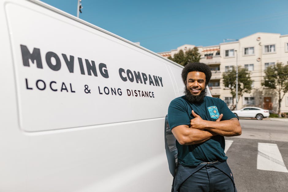 A smiling male removal driver with curly black hair and a beard, standing outdoors next to a white van branded with 'MOVING COMPANY LOCAL & LONG DISTANCE' in bold black letters, with his arms crossed. The van is parked on a street with multi-storey residential buildings in the background, and the sky is clear and blue. The driver is wearing a dark teal T-shirt with a small logo on the chest, and black trousers. Behind him, the pavement and a crosswalk are visible, indicating the loading area for home relocation or furniture transport. Inside the vehicle, cardboard boxes, wrapped furniture pieces, and packing materials like plastic wrap and blankets are visible through the open sliding door, suggesting an active packing and loading process. The scene reflects a professional moving service, such as those offered by Man With a Van Belmont, preparing for a local or long-distance move, with items staged for loading onto the van during a home relocation.