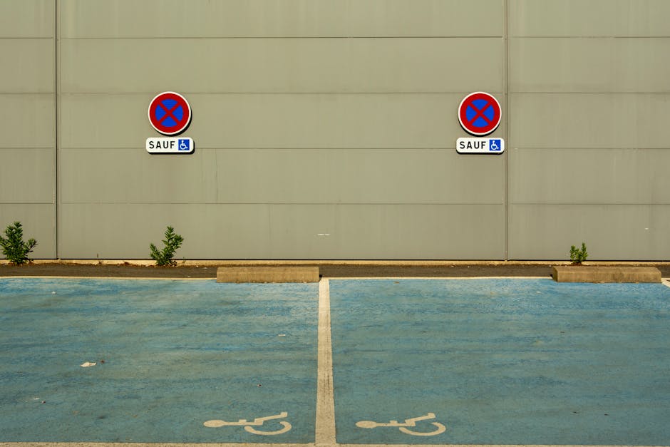 A photograph of a parking area adjacent to a plain, beige-colored wall. In the foreground, there are two blue handicap parking spaces marked with white wheelchair symbols on the ground. The parking spaces are separated by a white line placed centrally between them. Behind the spaces, there are two no parking signs mounted on the wall, each featuring a red circle with a blue background and a red cross, indicating no parking zones, with additional small signs below indicating exceptions for disabled badge holders. The ground is asphalt with some small debris, and there are small green shrubs growing along the base of the wall. The image captures a quiet, designated parking area, with subtle signage supporting parking rules relevant for moving and relocation logistics. Occasionally, details like the signs and markings may relate to helpful parking regulations during home relocation or furniture transport activities. The overall scene emphasizes space organization and regulations to facilitate effective vehicle and furniture movement in the area, with lighting indicating daylight hours.