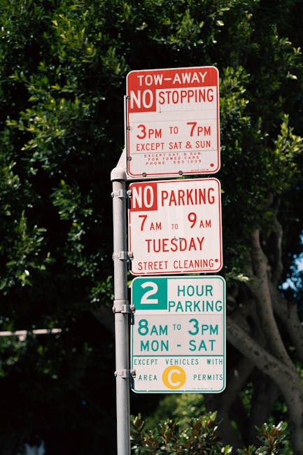 A tall metal street sign post in an outdoor setting displaying three different parking and stopping restrictions. The top sign is red and white, indicating a tow-away zone with no stopping permitted from 3 pm to 7 pm, except on Saturdays and Sundays, with a contact number for more information. The middle sign also red and white states no parking allowed from 7 am to 9 am on Tuesdays due to street cleaning. The bottom sign, in green, blue, and yellow, shows a 2-hour parking limit from 8 am to 3 pm Monday through Saturday, with an exception for vehicles displaying area C permits. The background features lush green foliage and partly cloudy sky, suggesting a suburban street corner, relevant to residential relocations and furniture transport logistics managed by Man With a Van Belmont.