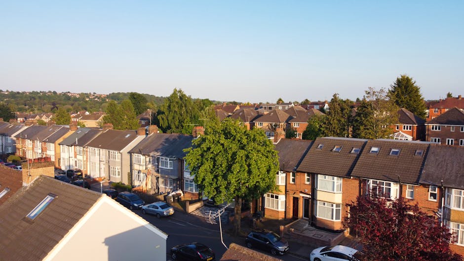 An aerial view of a residential street showing a row of terraced houses with pitched roofs, some with skylights, and multiple parked cars along the curb. Prominently visible in the foreground is the rooftop of a white house with a sky vent, casting a shadow on its roof. Several trees, including a large leafy green one and a smaller reddish-leaved tree, line the street, adding greenery to the scene. In the background, additional houses and trees extend towards the horizon under a clear blue sky. This image captures the typical environment where house removals and furniture transport activities might take place, with visible aspects of the moving process such as vehicle parking and location context provided by the street scene. It supports understanding of residential relocation logistics for services like those offered by Man With a Van Belmont, aligned with the page about Sutton Council permits for Belmont removals.