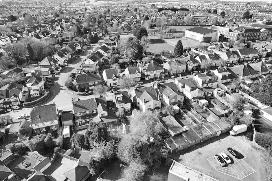 Black and white aerial photograph of a suburban residential area showing multiple detached and semi-detached houses with sloped roofs, gardens, and driveways. Some houses have visible vehicles parked outside, and a few are in the process of moving, indicated by covered furniture and open garage doors. In the foreground, a van from Man With a Van Belmont is parked on the pavement near several boxes wrapped in protective plastic and cardboard, arranged on a driveway and garden path. The loading process appears ongoing, with some furniture and boxes being prepared for transport. The image captures the urban landscape with neatly arranged houses, trees, and a sports field in the background, highlighting aspects of home relocation and furniture transport within the neighbourhood, consistent with professional house removals services related to the Sutton Council Permits process explained on the webpage.