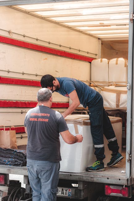 Two men are inside the open back of a moving truck, which is lined with a wooden roof and red horizontal support beams along the walls. One man, wearing a dark grey t-shirt with red text and grey trousers, stands on the ground near the truck, facing away from the camera. The other man, dressed in a blue t-shirt, black trousers, and sneakers with yellow accents, is standing on the truck bed, leaning forward slightly as he helps to lift or position a large white household object wrapped in protective plastic or padding. Surrounding items inside the truck include boxed cardboard packages, some wrapped with foam or bubble wrap, and various packing materials. The scene depicts typical packing and loading activities during a residential home relocation, with careful handling of furniture and packed belongings as part of a professional removals service, such as Man With a Van Belmont.