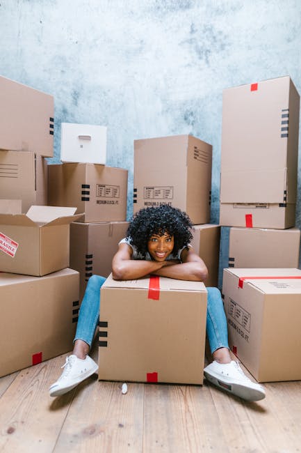 A young woman with curly black hair is sitting on the wooden floor inside a room, surrounded by numerous cardboard moving boxes of various sizes, some sealed with red or black tape and others partially open. The boxes are stacked against light blue textured walls, creating a scene of house packing preparatory to a home relocation. She is smiling and resting her chin on a medium-sized box in front of her, wearing casual white sneakers and blue jeans. The room appears to be filled with packing materials, and the scene suggests the process of loading or organizing items for transport, indicative of a professional furniture transport or removals service. This image is relevant to the context of house removals and moving logistics, closely aligning with the services provided by Man With a Van Belmont.