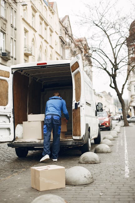 Two men are inside the open back of a moving truck, which is lined with a wooden roof and red horizontal support beams along the walls. One man, wearing a dark grey t-shirt with red text and grey trousers, stands on the ground near the truck, facing away from the camera. The other man, dressed in a blue t-shirt, black trousers, and sneakers with yellow accents, is standing on the truck bed, leaning forward slightly as he helps to lift or position a large white household object wrapped in protective plastic or padding. Surrounding items inside the truck include boxed cardboard packages, some wrapped with foam or bubble wrap, and various packing materials. The scene depicts typical packing and loading activities during a residential home relocation, with careful handling of furniture and packed belongings as part of a professional removals service, such as Man With a Van Belmont.
